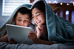 Two kids holding digital tablet while lying under blanket in the dark at night reading online book, watching or playing game before sleeping.  Little boy and sister lying in bed and faces illuminated by device screen light