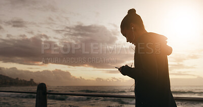Buy stock photo Woman, fitness and phone with earphones on beach for music, listening or sports podcast in nature. Active, female person or runner with mobile smartphone for sunset run, audio streaming or fit app