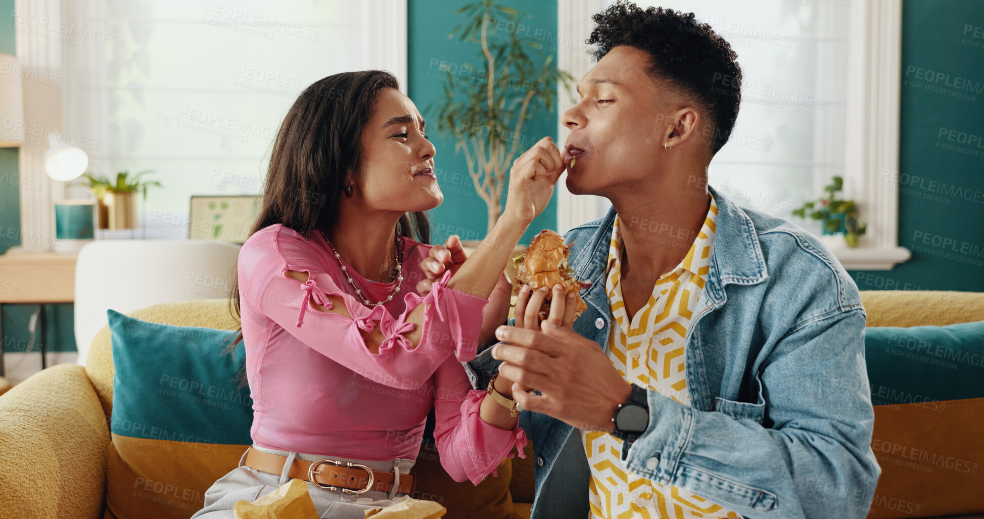 Buy stock photo Feeding, couple and delicious food for woman, home or people with meal in living room, smile or takeaway. House, happy and man in relationship with hungry partner enjoying taste of burger in lounge