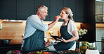 Cooking, mature couple and feeding in kitchen, eating and bonding with ingredients for cuisine in home. Healthy food, helping and people with tablet for research of recipe, culinary skills or house