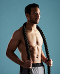 Handsome young hispanic man training with battle ropes in studio isolated against a blue background. Mixed race shirtless male athlete exercising or working out to increase his strength and fitness