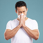 Handsome young mixed race man blowing his nose while standing in studio isolated against a blue background. Hispanic male suffering from cold, flu, sinus, hayfever or corona and using a facial tissue