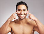 Handsome young mixed race man shirtless in studio isolated against a grey background. Hispanic male applying moisturizer lotion to his face. Looking confident and happy with his daily skincare regime