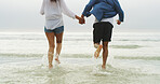 Couple, holding hands and running in water at beach for travel holiday, freedom or support in relationship. Back, legs of people and romantic date by ocean with adventure, trust or honeymoon vacation