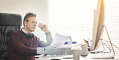 Buy stock photo A businessman analysing paperwork and using a computer in a modern office. A mature male executive working online and going over a business document at his desk