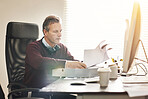 A businessman analysing paperwork and using a computer in a modern office. A mature male executive working online and going over a business document at his desk