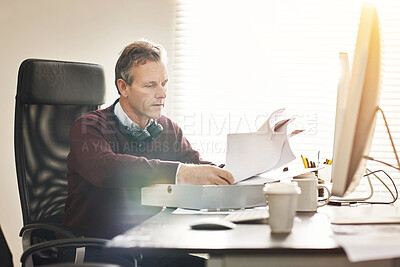 Buy stock photo A businessman analysing paperwork and using a computer in a modern office. A mature male executive working online and going over a business document at his desk