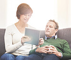 A mature couple using a digital tablet, relaxing at home. A happy senior man and woman in love browsing online with a touchscreen on the couch