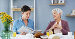 Senior woman, caregiver and reading book in home, breakfast or smile in retirement in dining room. People, connection and literature in morning for care, storytelling and support with novel in house