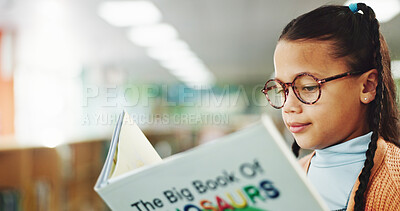 Buy stock photo Girl, child and reading book at library for education, learning or concentration with dinosaur story. School kid, glasses and scholarship with language, knowledge and study for development at academy
