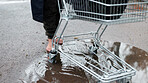 Homeless, person and walking with shopping cart in street for poverty, economic hardship or vulnerability. Barefoot, beggar and trolley in rain puddle for welfare crisis, struggle or social injustice