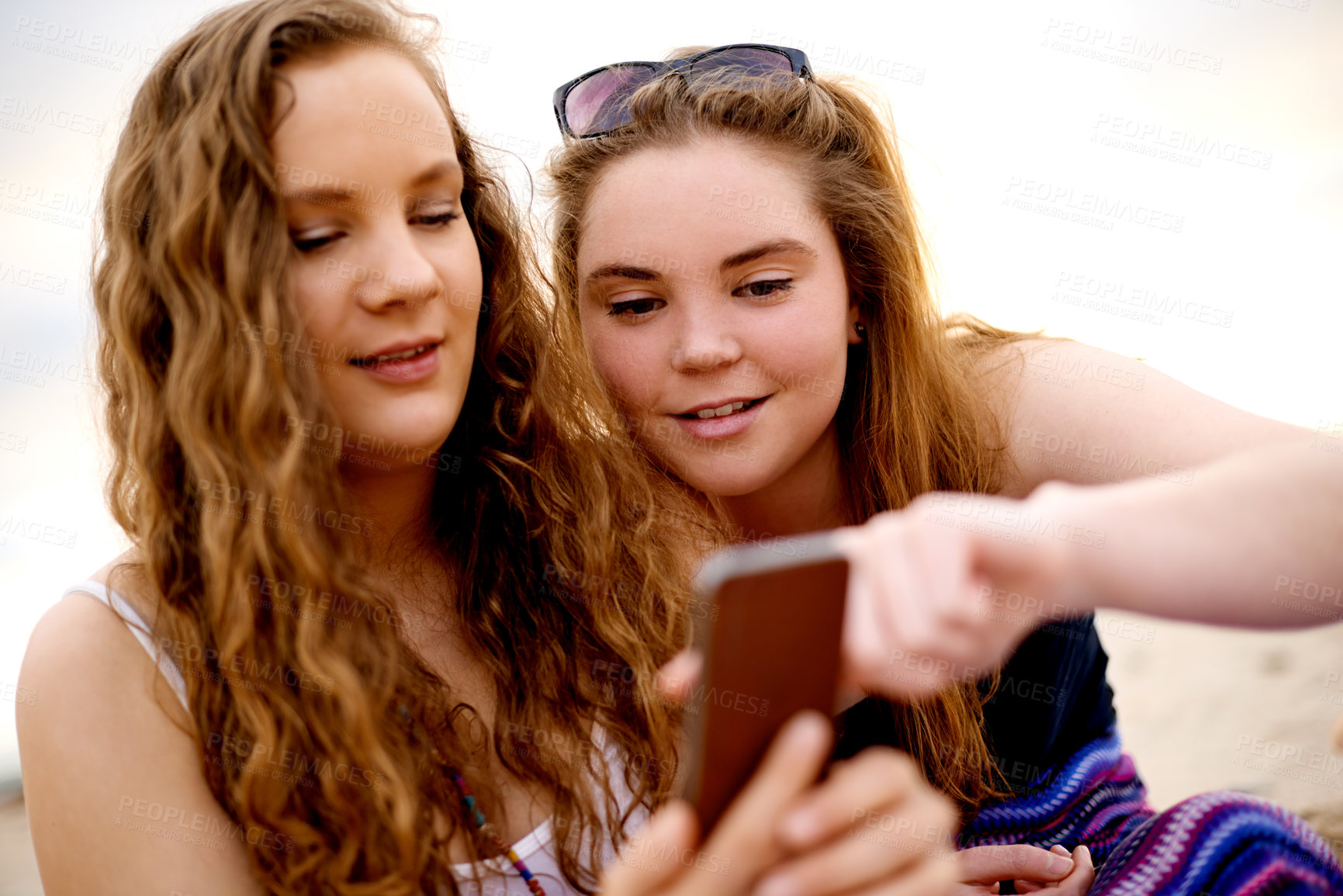Buy stock photo Girl, friends and phone at beach, texting and happy with meme, post and scroll with chat on vacation in summer. People, smartphone and search on web, mobile app or social media at sunset in Spain
