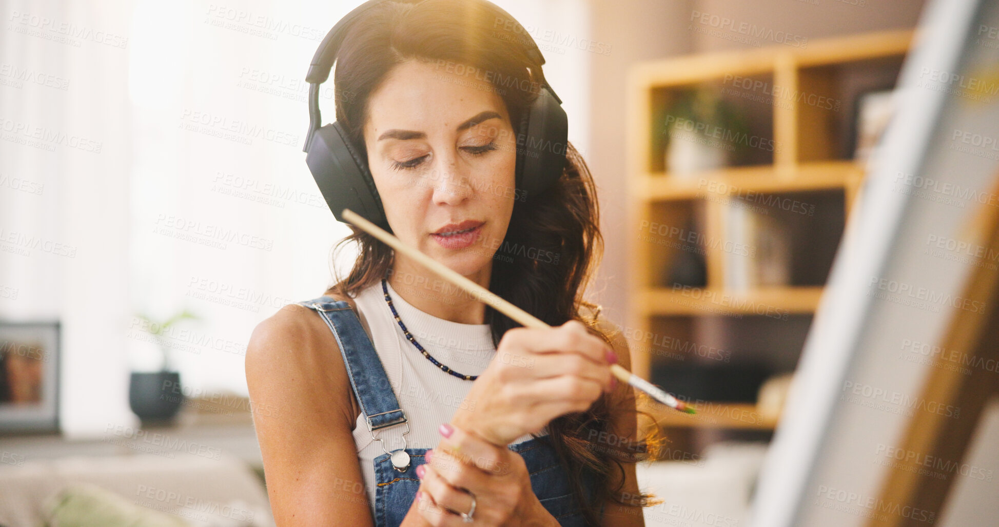 Buy stock photo Music, paint brush and wrist pain with woman in living room of home for artistic or creative hobby. Arthritis, carpal tunnel or headphones and mature painter in apartment with canvas on easel