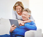 Happy mature woman hugging her husband from behind and watching him browse online on a digital tablet while relaxing together at home