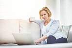 Mature woman drinking coffee and browsing online on a laptop while relaxing alone on a sofa at home