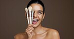 Beauty, woman and happy with makeup brushes in studio to apply cosmetics, skin glow and foundation. Mockup space, excited and girl with skincare product for concealer or self care on brown background