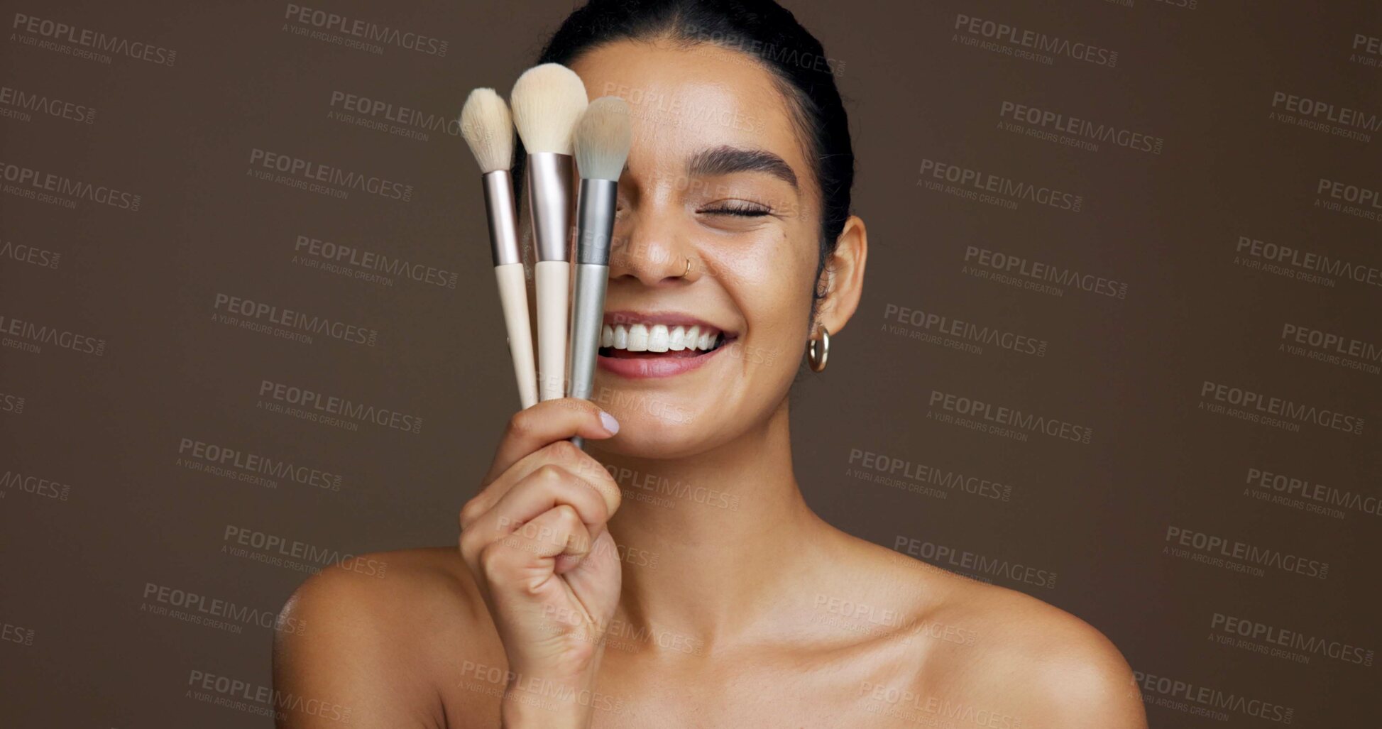 Buy stock photo Beauty, face and makeup brush selection with woman in studio for cosmetics or makeover tools. Equipment, facial and smile of happy model person on brown background to apply concealer or foundation