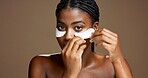 Black woman, apply and eye mask in studio for skincare, portrait or reduce puffiness. Model, brown background and hyaluronic patches for hydration, beauty glow or cosmetic product for dark circles