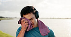 Tired man, sweat or fitness with towel or headphones at lake for intense workout, break or recovery. Active, male person or dry with cloth for physical activity, exercise or outdoor training at river