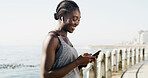 Happy, black woman and fitness with phone on beach promenade for sport app or exercise schedule. Active, female person or smile with mobile smartphone, earphones or listening to music on ocean coast