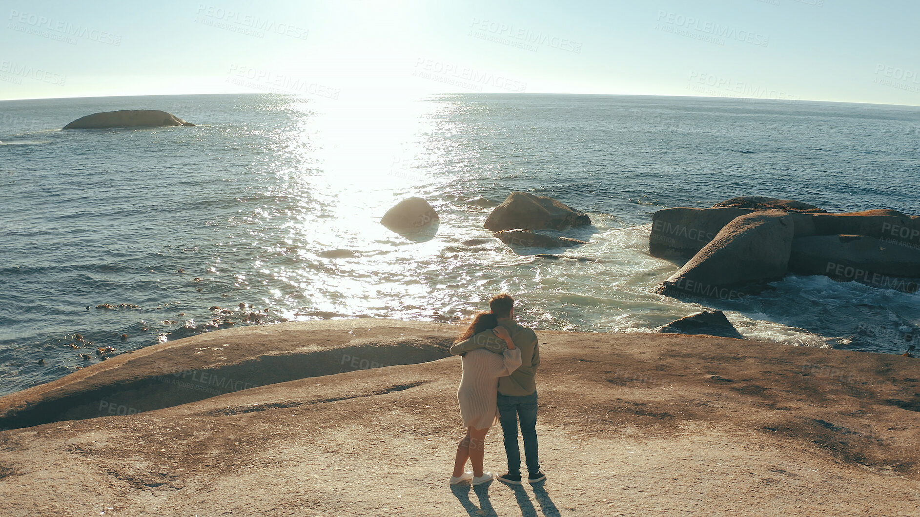 Buy stock photo Hug, sunrise and back of couple by ocean for relaxing on holiday, vacation and weekend together. Dating, above and man with woman embrace by sea, water and rocks for bonding, relationship or romance