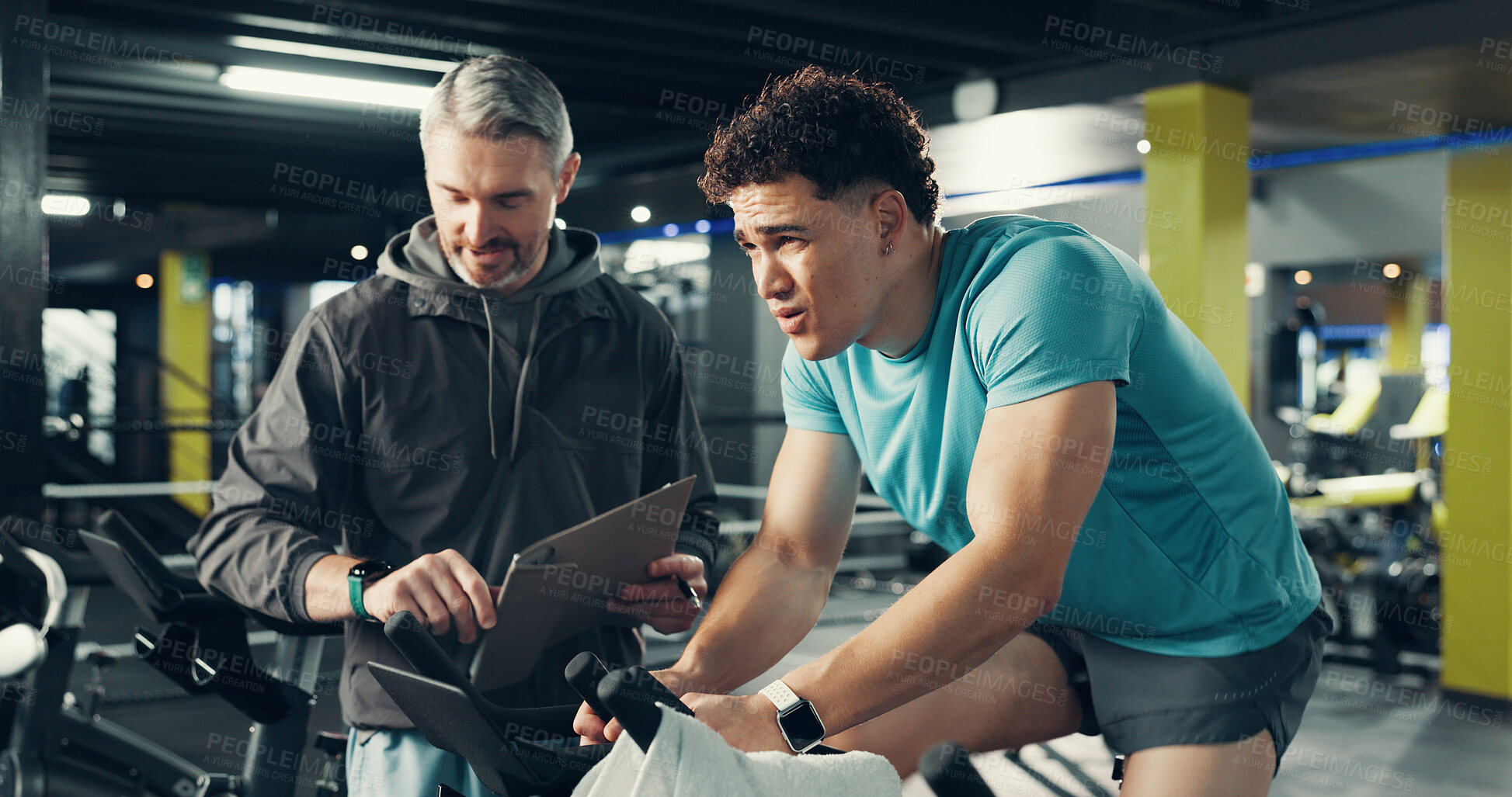 Buy stock photo Stationary bicycle, man and coach for fitness, help or workout timer for health in gym. Cycling, personal trainer and training on bike for spin class, challenge or check watch for exercise progress