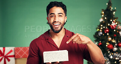 Buy stock photo Portrait, gift box and man in studio for christmas, festive or tradition celebration with pointing. Happy surprise, holiday and male person with present by xmas tree with pride by green background.