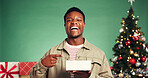 Portrait, laughing and man with present, Christmas or excited for festive season, smile and pointing. Studio, black person and Xmas celebration with gift, green background and tree with ornaments