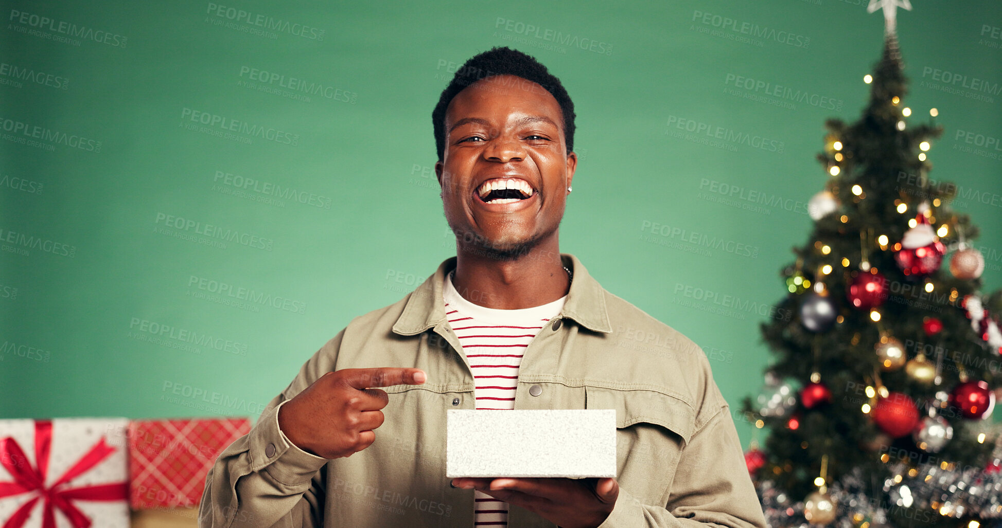 Buy stock photo Portrait, laughing and man with present, Christmas or excited for festive season, smile and pointing. Studio, black person and Xmas celebration with gift, green background and tree with ornaments