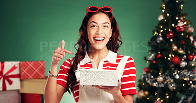 Buy stock photo Portrait, gift box and woman in studio for christmas, festive or tradition celebration with pointing. Happy surprise, holiday and person with present by xmas tree with pride by green background.