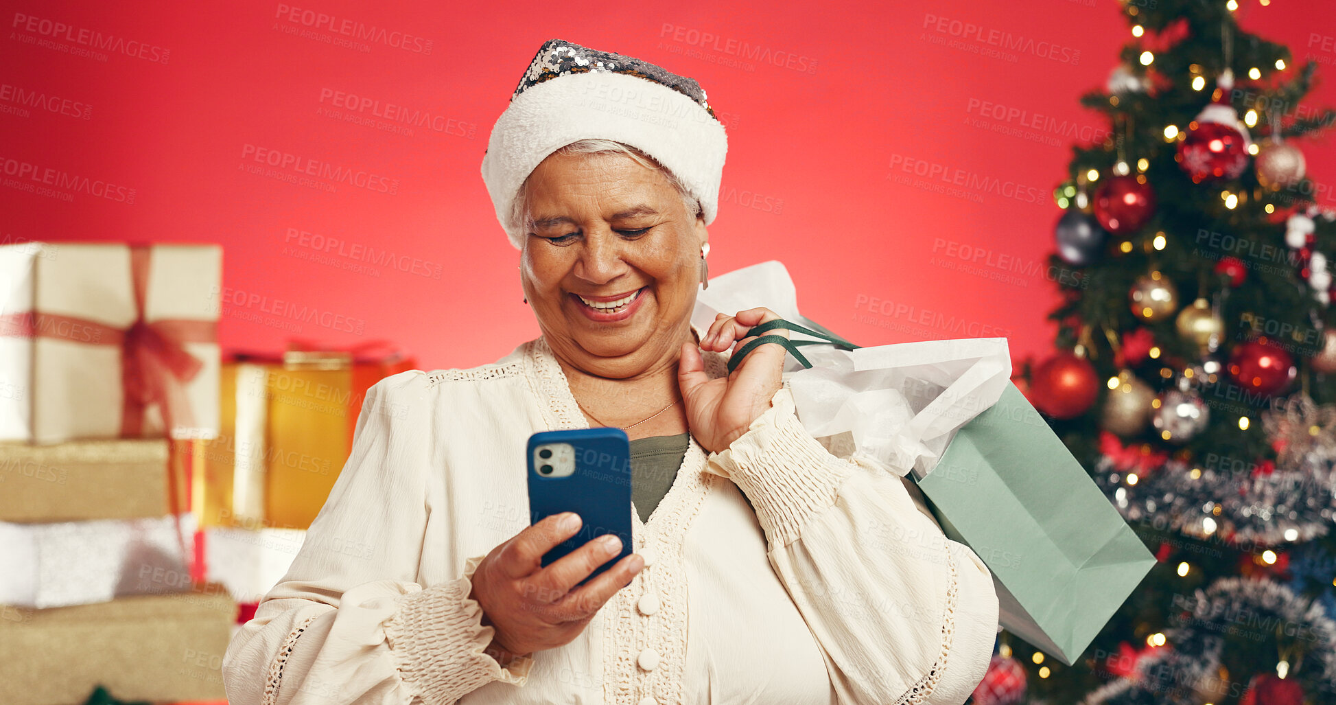 Buy stock photo Cellphone, shopping bags and senior woman with christmas in studio with networking on mobile app. Laugh, gifts and elderly female person with phone for communication with xmas by red background.