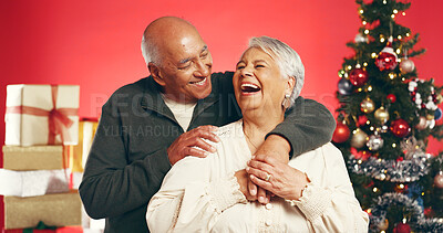 Buy stock photo Hug, laugh and senior couple with Christmas in studio with love, festive and marriage tradition. Happy, retirement and elderly man embracing woman by xmas tree for celebration on red background.