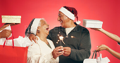 Buy stock photo Senior couple, hug and christmas with fireworks for festive celebration on a red studio background. Elderly, man or woman with smile, sparkler or shopping bags for gifts or december holiday together