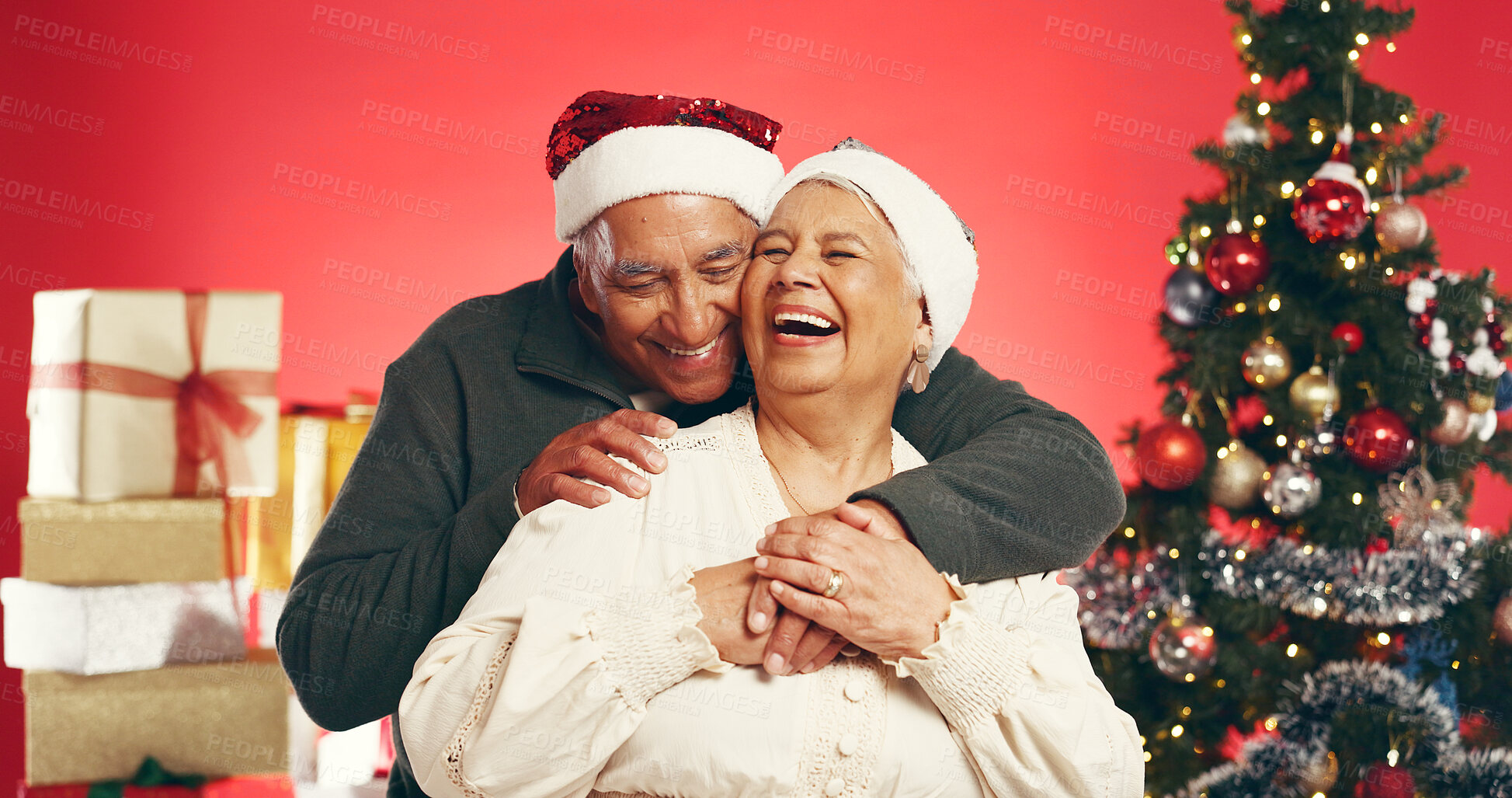 Buy stock photo Laugh, christmas and senior couple in studio with love, festive and marriage tradition. Hugging, santa hat and elderly man embracing woman by xmas tree for celebration together by red background.