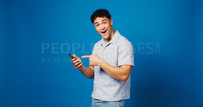 Buy stock photo Man, surprise and pointing at phone in studio with news, notification or sale on blue background. Wow, mobile and portrait of happy person with shock for discount, promo or giveaway with mockup space
