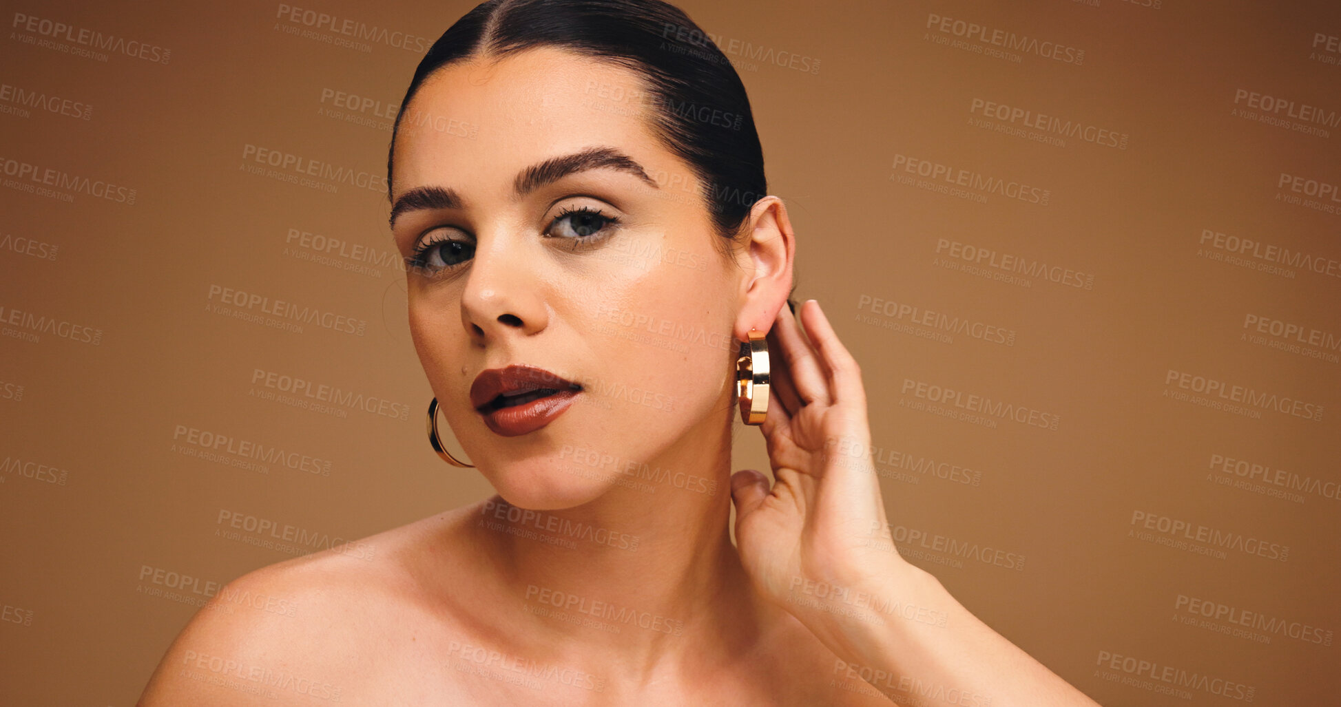 Buy stock photo Earrings, beauty and woman with makeup in studio for aesthetic, glow and shine on brown background. Ear jewelry, portrait and model with lipstick cosmetics, face transformation and confidence