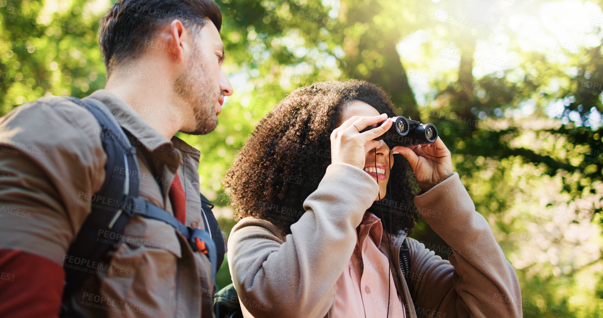 Buy stock photo Couple, hiking and happy with binoculars in forest for sightseeing, adventure and outdoor in summer. Woman, man and interracial love for birdwatching, direction or smile with trekking in woods
