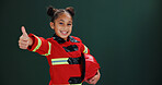 Happy girl, portrait and firefighter with thumbs up in studio for career day on a green background. Child, kid or smile with like emoji, yes sign or OK in costume for safety success on mockup space