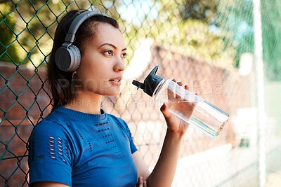 Buy stock photo Sports, woman and drinking water with headphones for music, fitness playlist or motivation with podcast. Fence, athlete and thinking with h2o for thirst, hydration or listen audio in basketball court