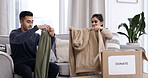 Couple, folding clothes and donation box in house for charity initiative, nonprofit or declutter home. Married people, apparel or giveaway in package for NGO contribution, support or spring cleaning.