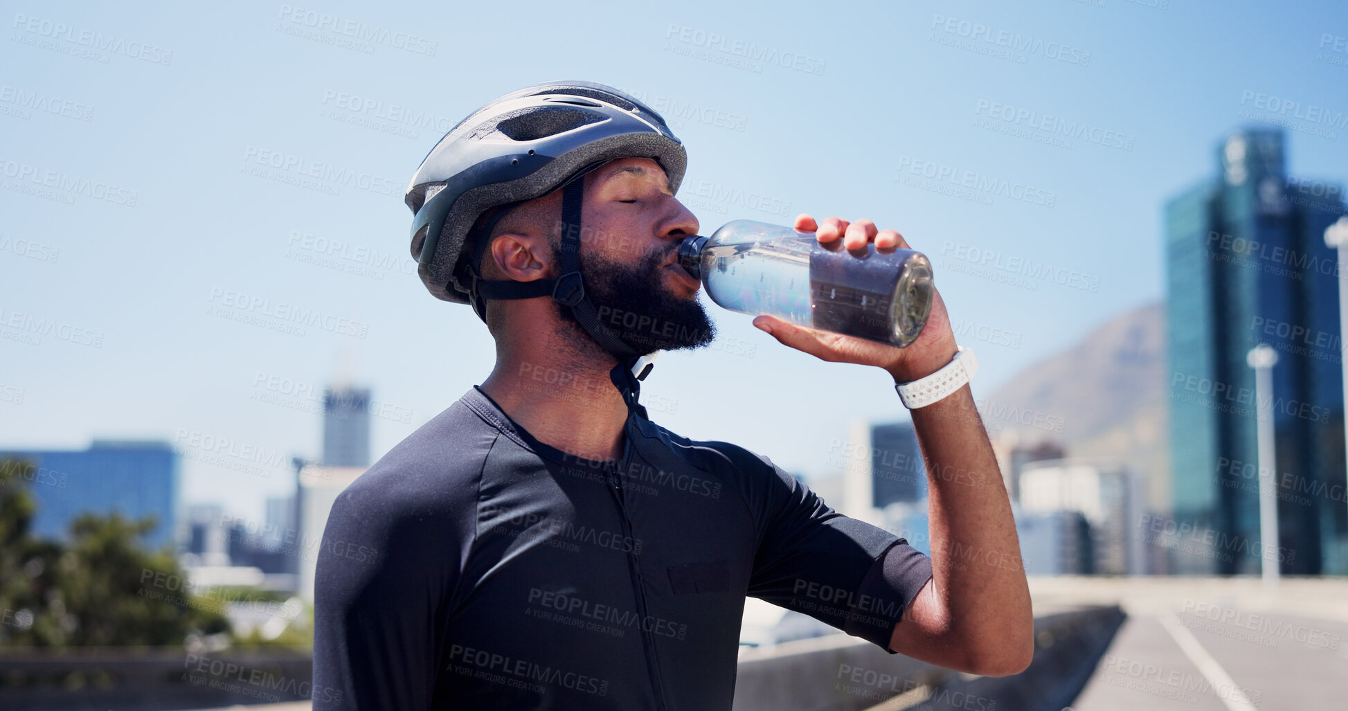 Buy stock photo Black man, cyclist and drinking water in city, fitness and helmet on bridge, break and hydration in street. African person, bottle and rest with training, outdoor and liquid for electrolytes in town