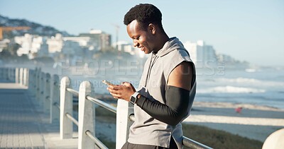 Buy stock photo Black man, phone and texting with fitness on beach promenade with smile, break and application. Person, runner and happy with smartphone, check notification and outdoor for exercise on boardwalk