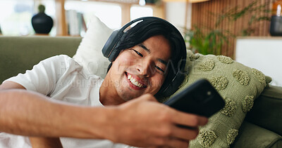 Buy stock photo Headphones, phone and man on sofa in home listening to music with texting, networking or social media. Happy, audio tech and Asian male person with cellphone for streaming playlist on mobile app.