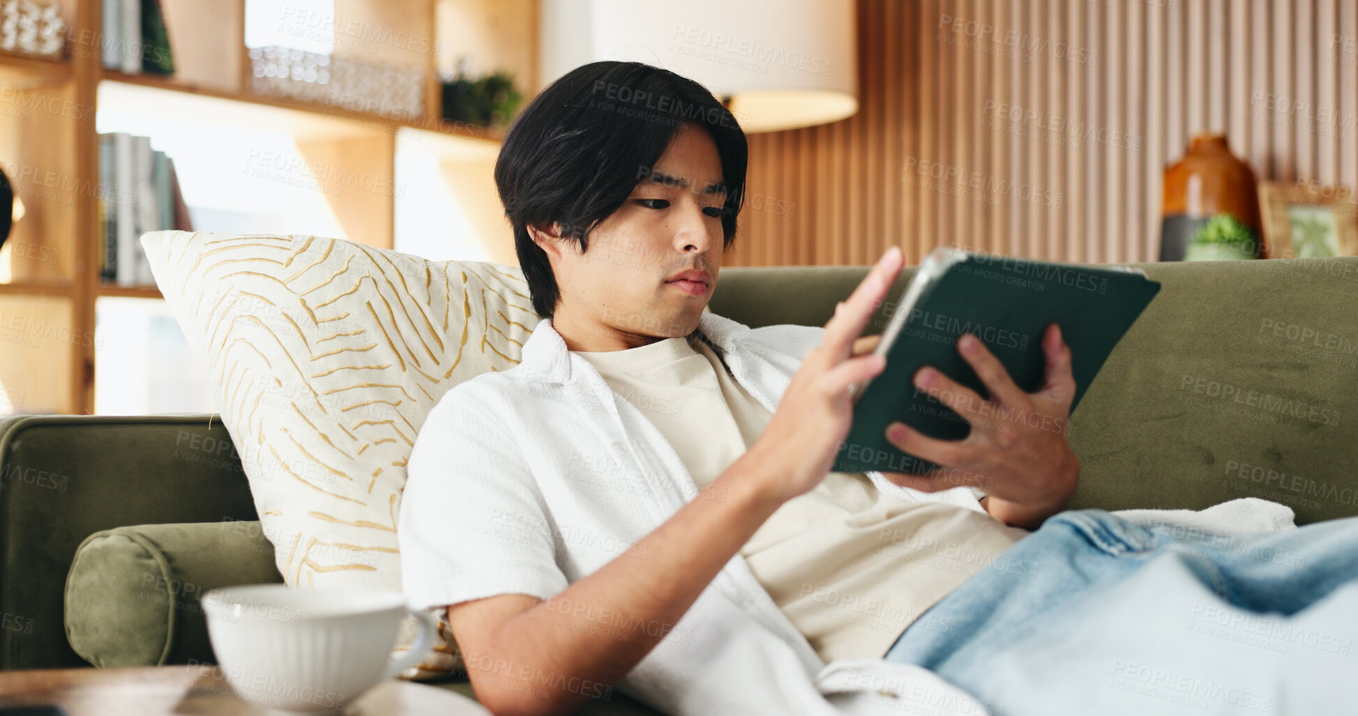 Buy stock photo Education, search and tablet with Asian man on sofa in home living room for distance learning. App, online study and streaming with serious university student in apartment for development or growth