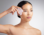 Studio closeup of a beautiful mixed race woman using a rose quartz gua sha to reduce wrinkles and puffiness. Young hispanic woman using anti ageing tool against grey copyspace background