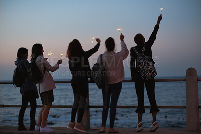 Buy stock photo Sunset, friends and sparkler by beach with new year celebration, bonding together and fun holiday. Back, evening and women outdoor on promenade with ocean, fireworks or travel adventure for vacation.