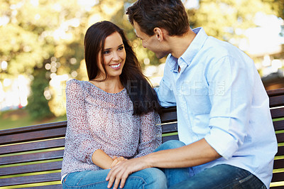 Buy stock photo Loving young couple talking happily on a park bench on a sunny afternoon