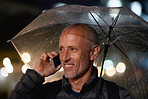 Mature man using smartphone having phone call talking on mobile phone holding umbrella in rainy city at night