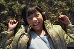 Funny little asian girl wearing headphones lying on grass smiling having fun listening to music in park