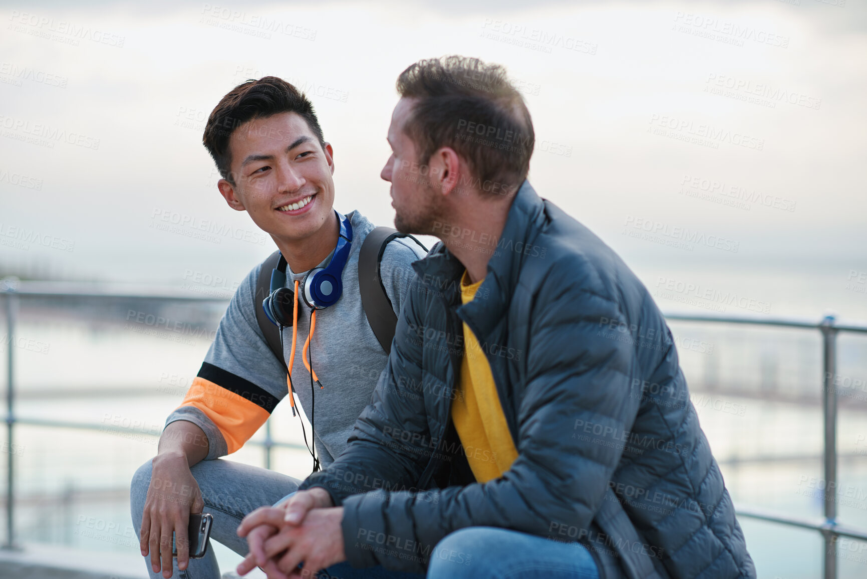 Buy stock photo Men, friends and chat on promenade at beach with smile, bonding and conversation on seaside vacation. People, relax and holiday with discussion, memory or tourism with break on getaway by ocean